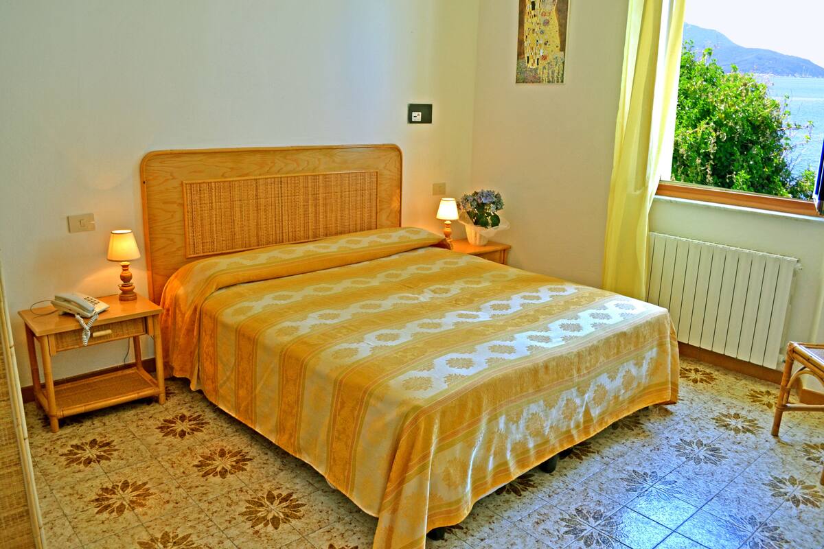 A bedroom with a bed, two bedside tables, a nightstand, a lamp, a potted plant, and a window with a view of greenery and mountains.