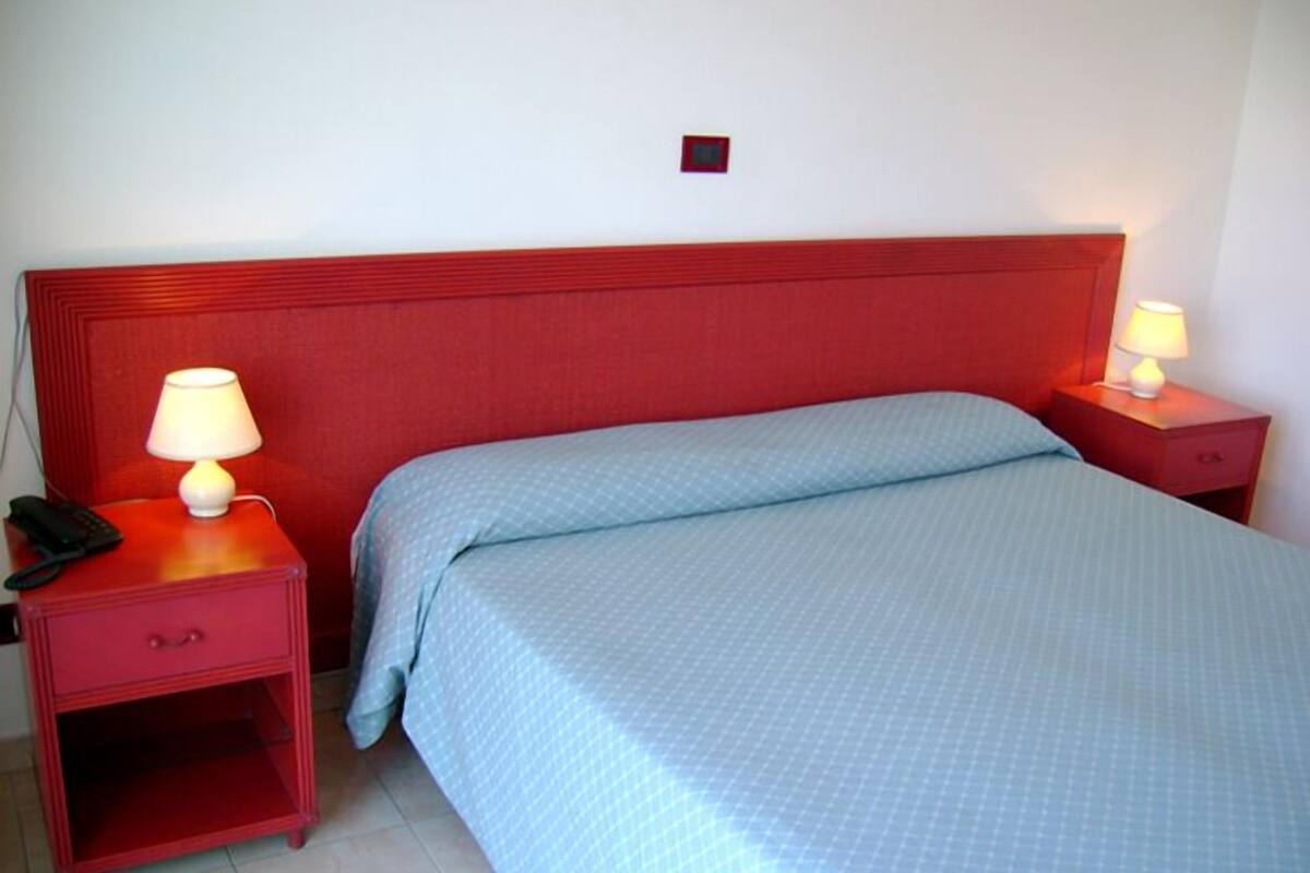 A hotel room with a red headboard, two bedside tables with lamps, a bed with a blue bedspread, and a red telephone on the left table.