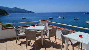 Terraço com mobiliário de vime e uma mesa, com vista para um mar calmo, barcos e montanhas ao fundo.
