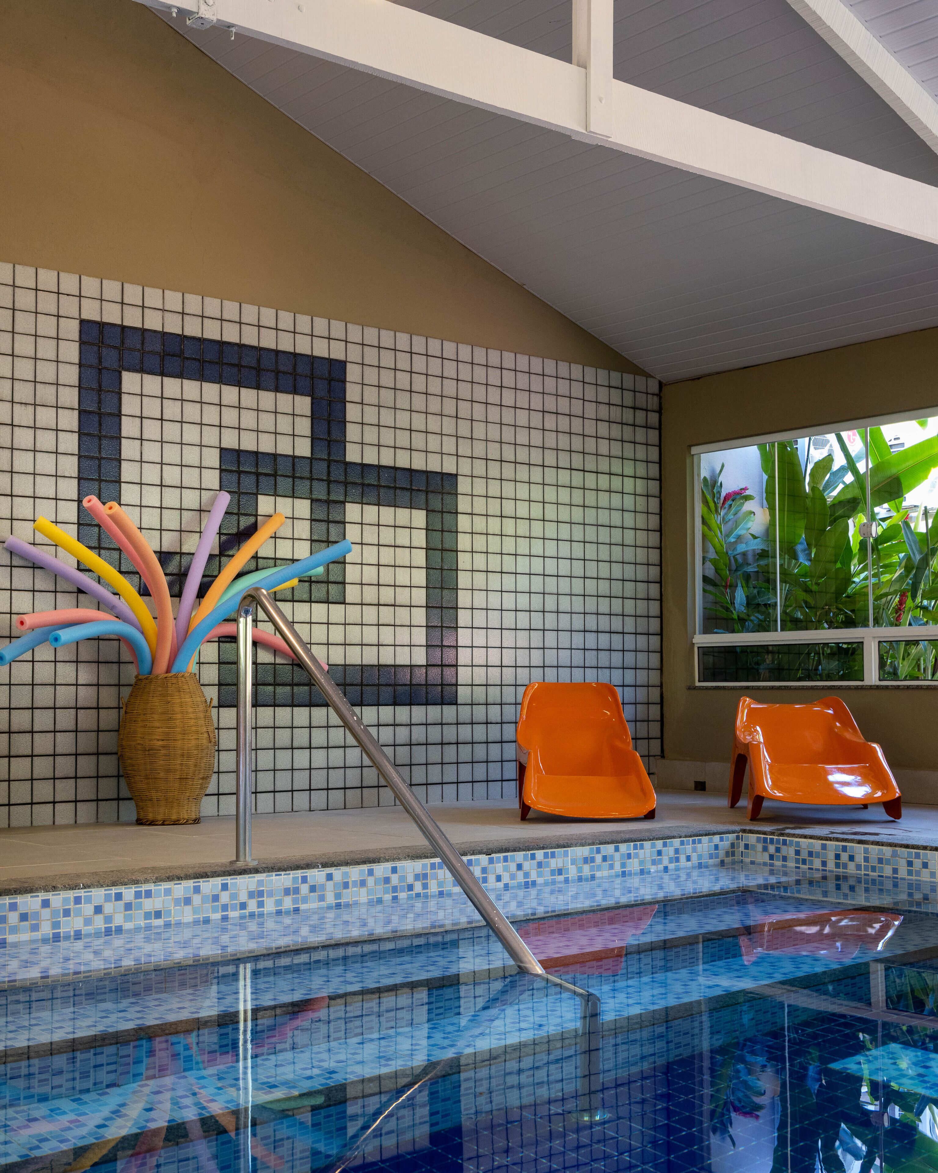 Uma área de piscina com paredes revestidas de azulejos, uma cesta de vime com objetos coloridos e duas cadeiras laranja.