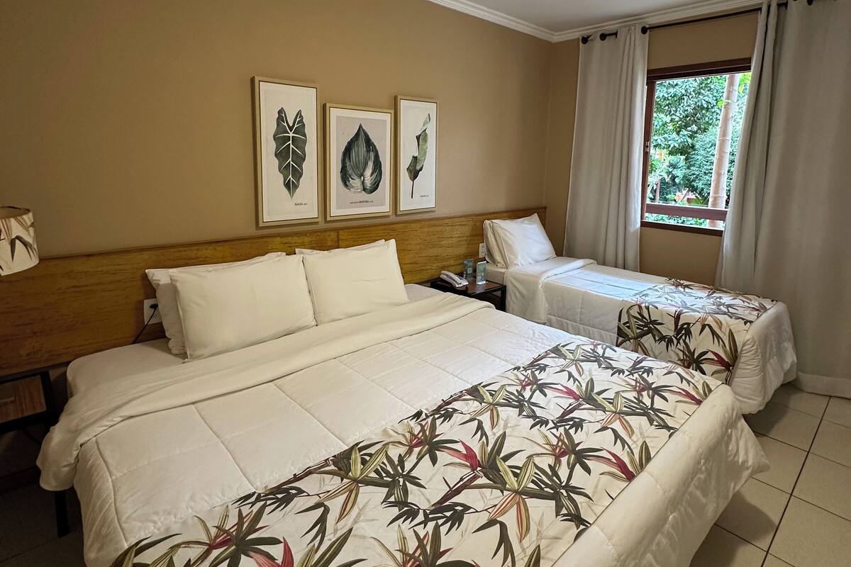 A hotel room with a large bed, a smaller bed, and a window with curtains. There are three framed botanical prints on the wall.