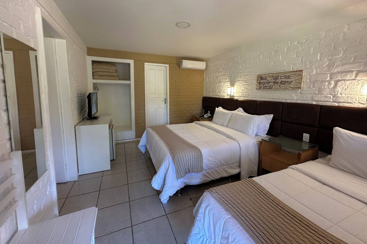 A hotel room with two beds, a wardrobe, a television, and a brick wall with a light fixture.
