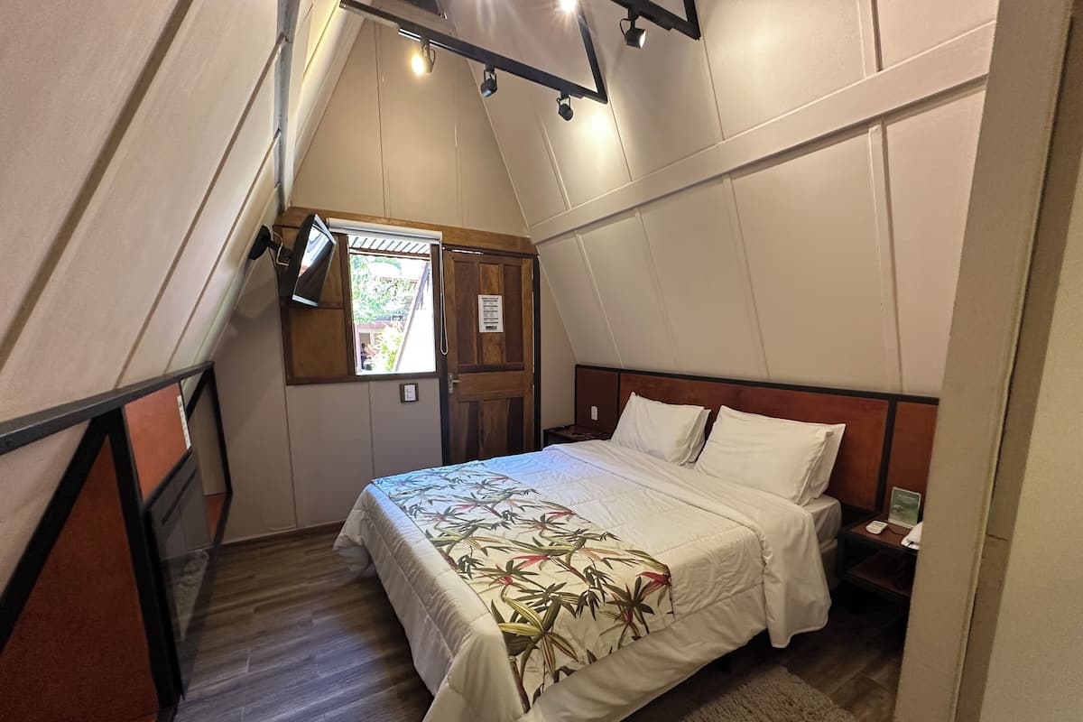 A cozy, well-lit attic bedroom with a wooden floor, a bed with a floral bedspread, a nightstand, and a window with a view of greenery outside.