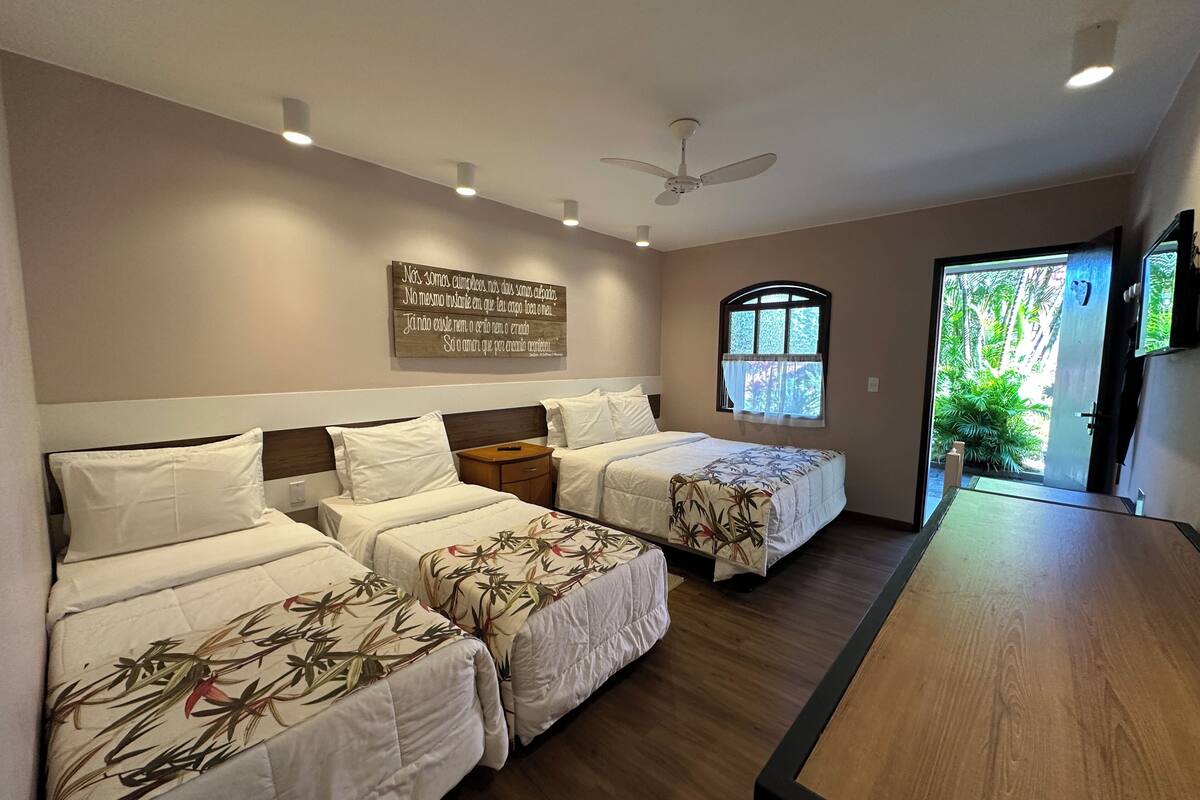 A hotel room with two beds, a wooden headboard, a ceiling fan, recessed lighting, a window with a view of greenery, and a wooden table.