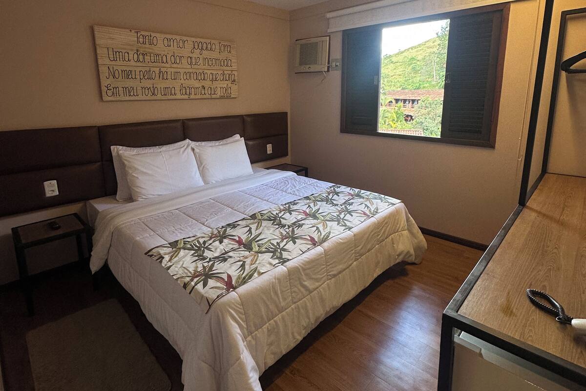 A bedroom with a bed, bedside tables, a wooden desk, a window with a view, and a wall-mounted air conditioner.