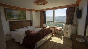 A hotel room with a bed, a desk, a chair, a mirror, and a view of the mountains. - Ajloun Hotel (Ajloun)