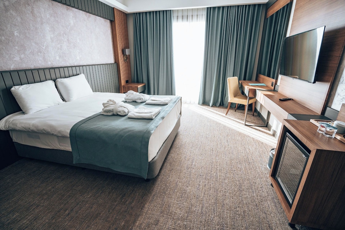 Habitación de hotel con cama, escritorio, silla, televisor y ventana con cortinas.