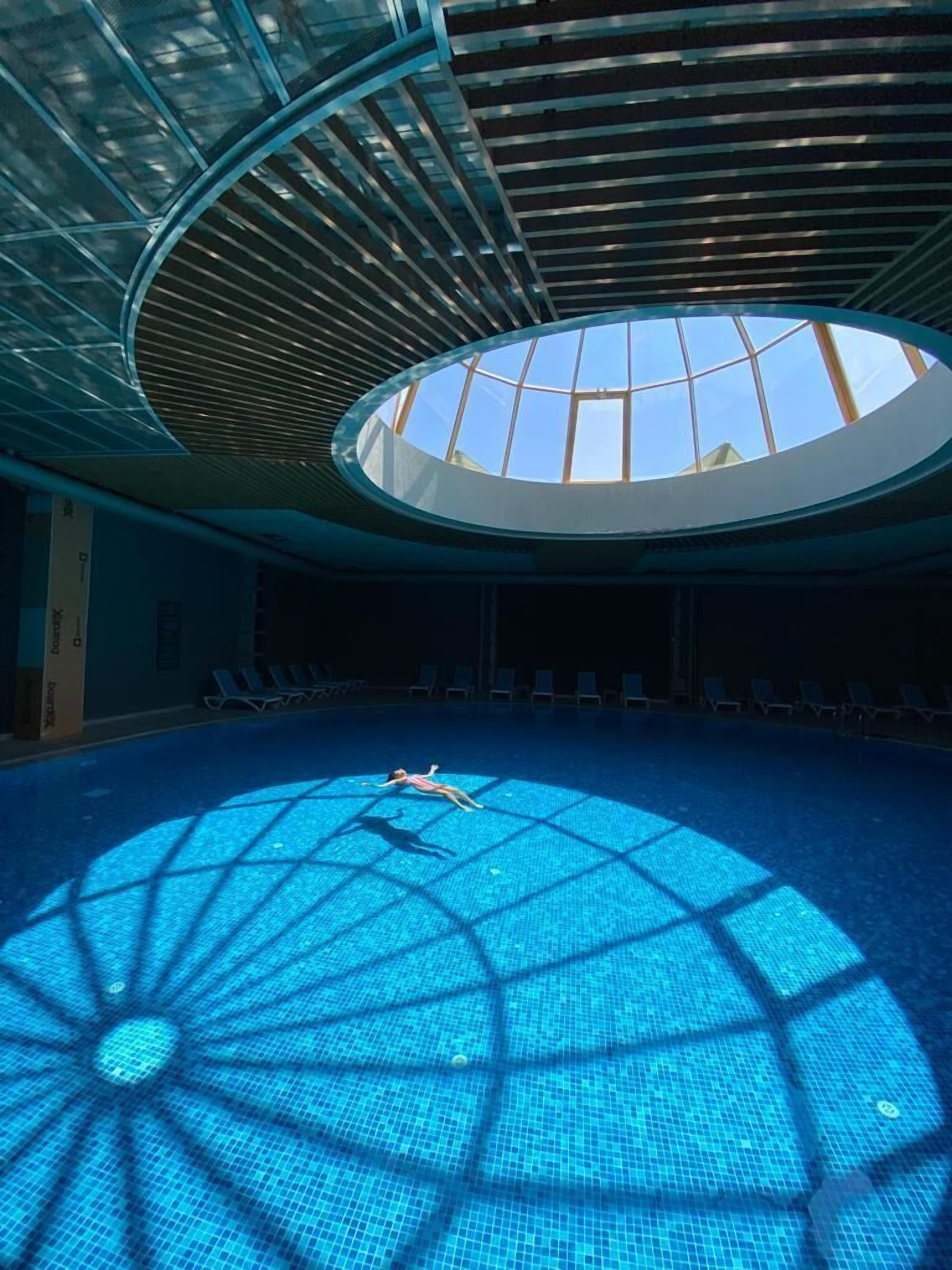 Una piscina interior con techo de tragaluces, agua azul cristalina y reflejos de sol en el suelo.