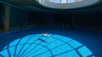 Una piscina interior con techo de tragaluces, agua azul cristalina y reflejos de sol en el suelo.