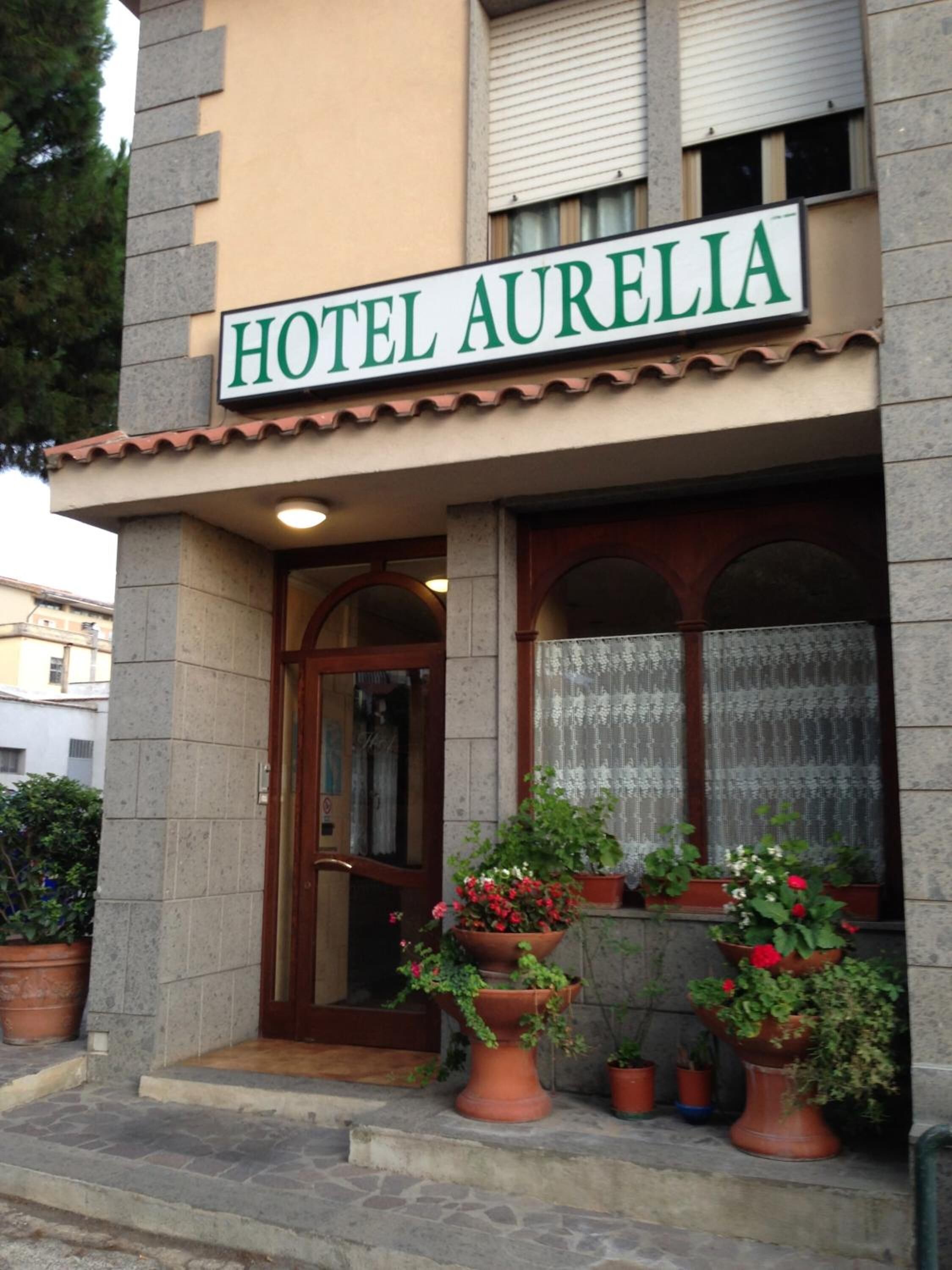 Ett hotell som heter HOTEL AURELIA, med en fasad av sten, takpannor i terrakotta och en inbjudande entré med trappsteg.