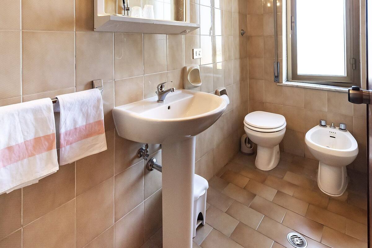 A bathroom with a sink, two toilets, a bidet, a mirror with lights, and a window.