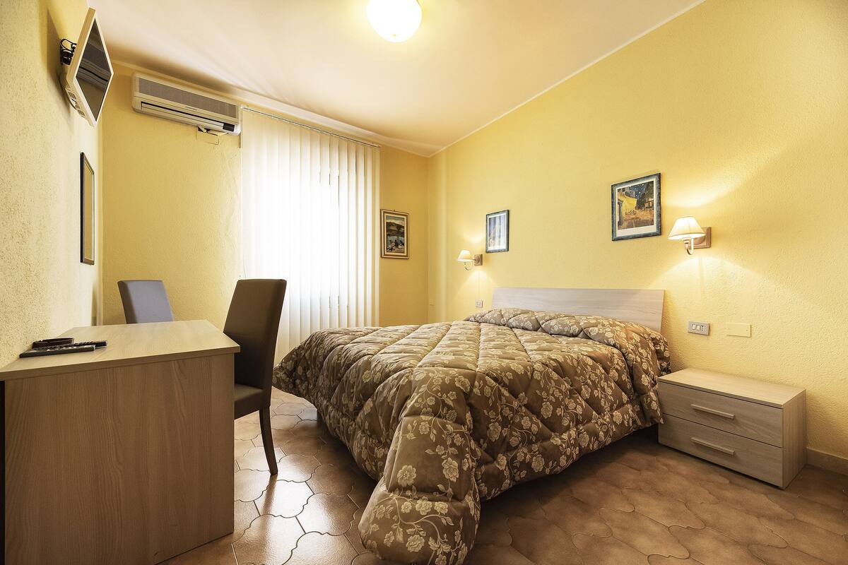 A hotel room with a bed, a desk, a chair, a nightstand, and two wall-mounted lamps.