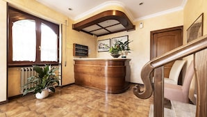 A reception area with a wooden reception desk, a potted plant, a radiator, and a staircase. - Hotel Aurelia (Tarquinia)