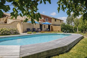 A swimming pool with a wooden deck, lounge chairs, and a building with a tiled roof in the background.