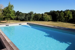 A rectangular swimming pool with a wooden deck edge, surrounded by a grassy area and trees.