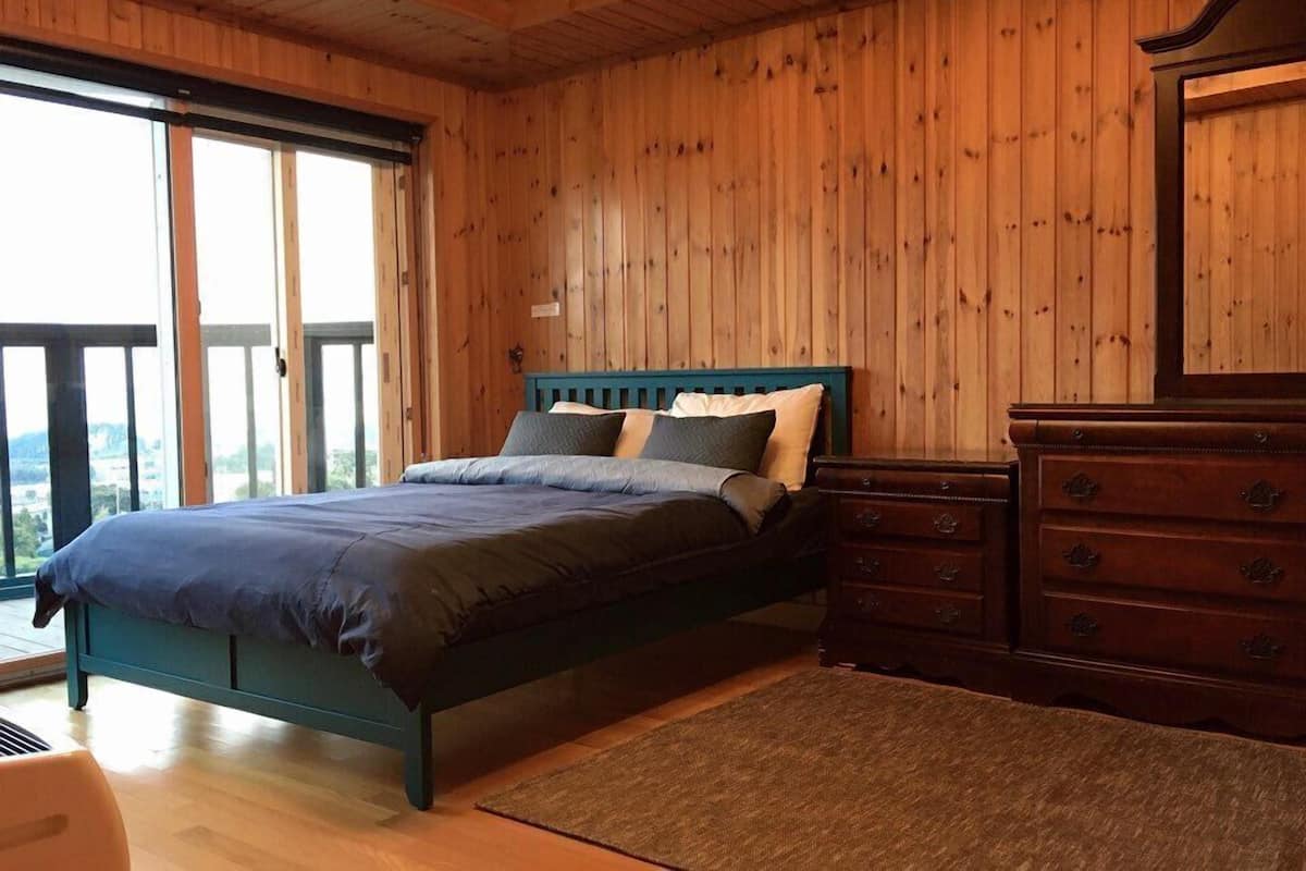 A bedroom with a bed, wooden walls, a dresser, and a view through a large window.