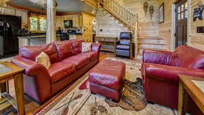 A spacious log cabin interior with a leather sofa, ottoman, and a chair. A staircase leads to an upper level. - Gorgeous 4BR Log Home Near Storyland & N. Conway. AC, Large Deck, Dog friendly (Bartlett)