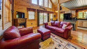 A spacious living room with a fireplace, leather sofas, and a large window overlooking a deck. - Gorgeous 4BR Log Home Near Storyland & N. Conway. AC, Large Deck, Dog friendly (Bartlett)
