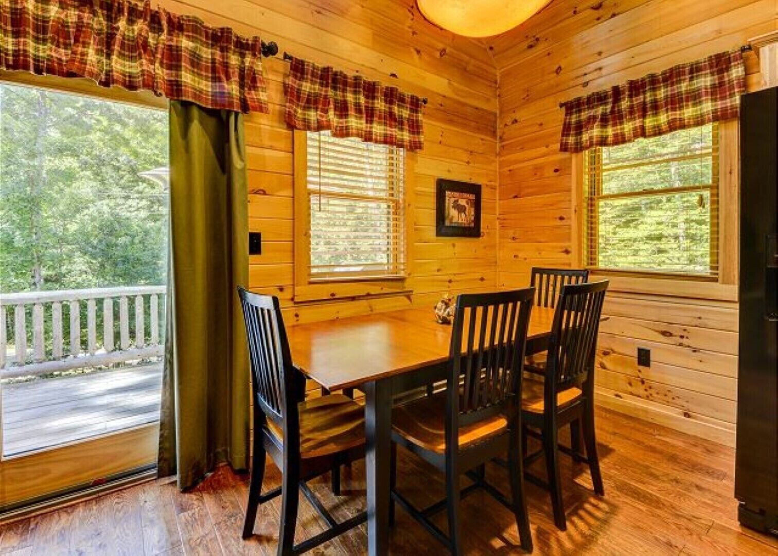 A wooden cabin interior with a dining table and four chairs, large windows with plaid curtains, and a view of a road and greenery outside.