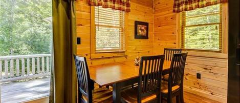 A wooden cabin interior with a dining table and four chairs, large windows with plaid curtains, and a view of a road and greenery outside.