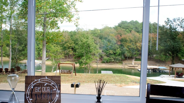 A table with a wooden sign reading "BE OUR Guests," a notebook, and a glass hourglass. Outside, there's a view of a lake, trees, and a dock with chairs and a swing. - 295 is 5 minutes from dam with boat launch and tritoon boat for rent. (Jasper)