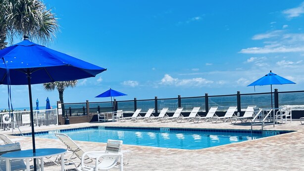 A pool area with white lounge chairs and blue umbrellas, a tiled deck, and a view of the ocean.