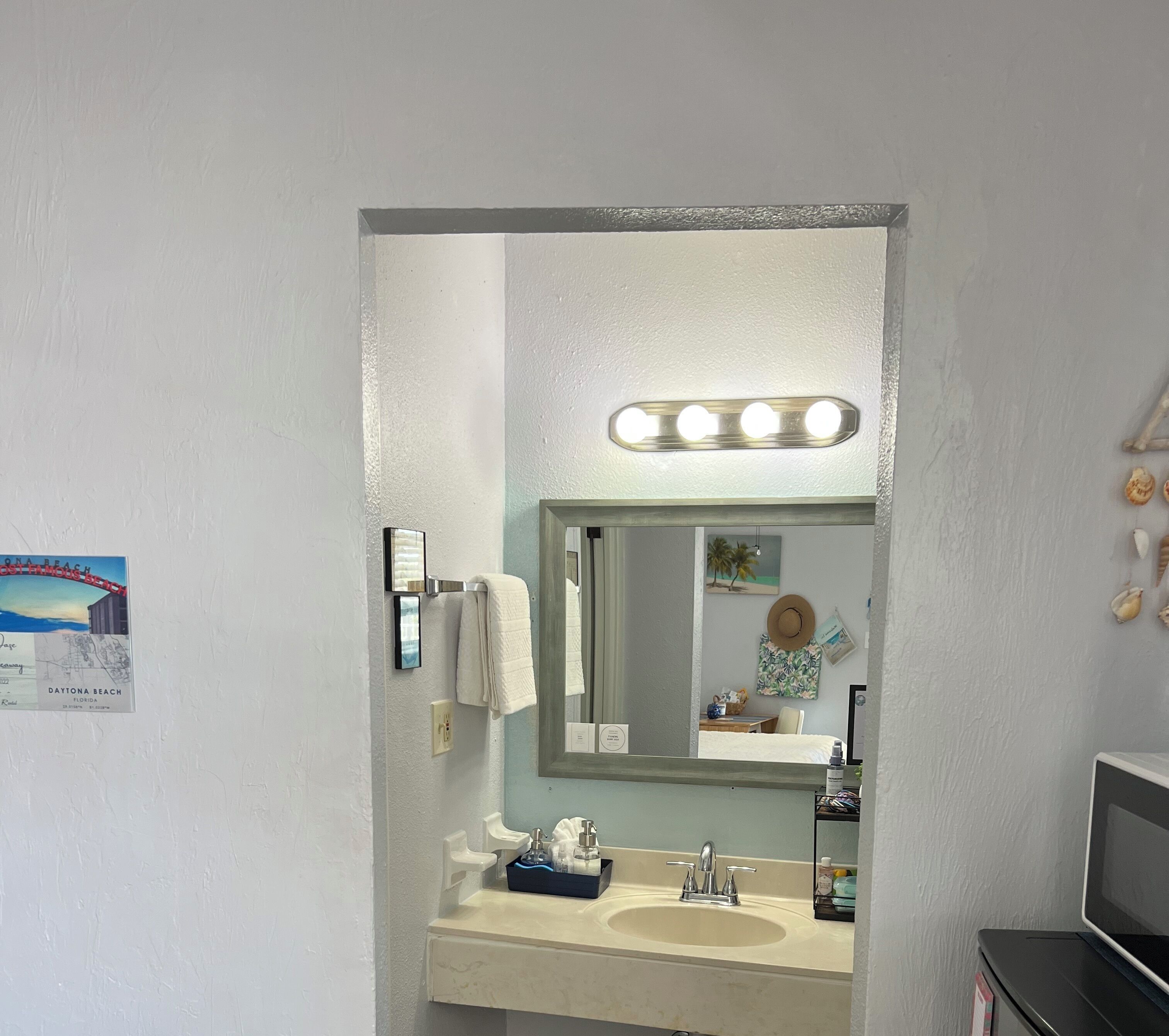 A bathroom with a sink, mirror, and vanity. There is a sign on the wall and a towel hanging on the door.
