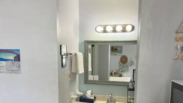 A bathroom with a sink, mirror, and vanity. There is a sign on the wall and a towel hanging on the door.