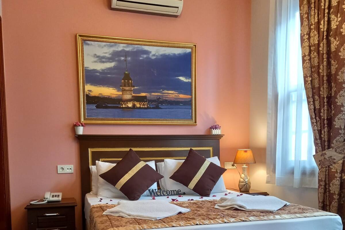 A hotel room with a bed, a nightstand, a bedside lamp, a window with curtains, and a painting above the bed.
