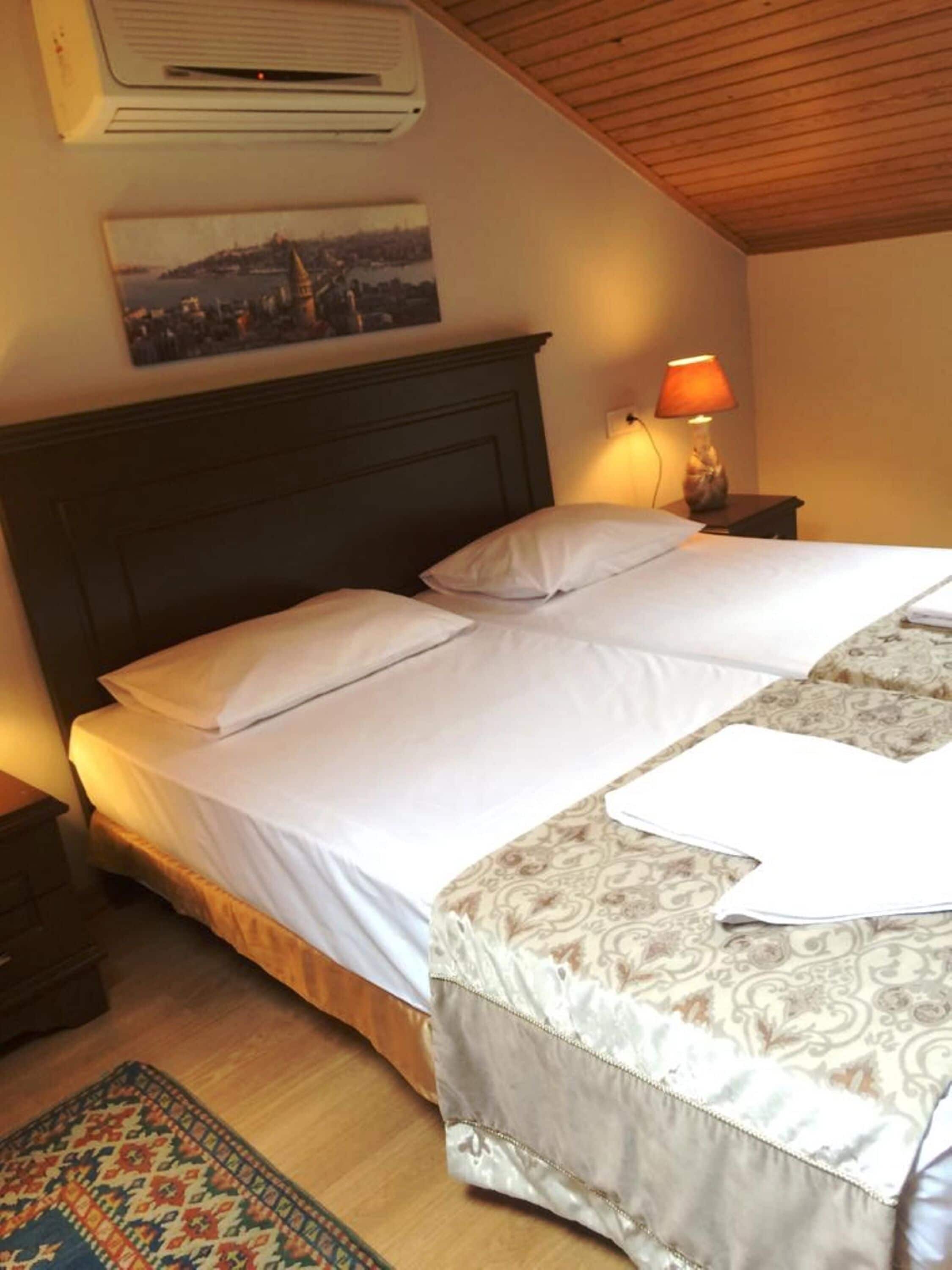 A hotel room with a large bed, a nightstand, a lamp, an air conditioner, and a patterned rug.