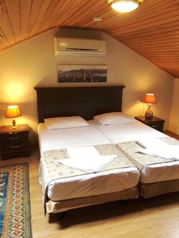 A cozy bedroom with a large bed, two bedside tables, a nightstand, a wooden ceiling, and an air conditioning unit.
