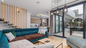 Een moderne woonkamer met een blauwe bank, een salontafel en een grijze fauteuil. Achtergrond is een keuken zichtbaar, en er is een grote schuifdeur die leidt naar een terras.