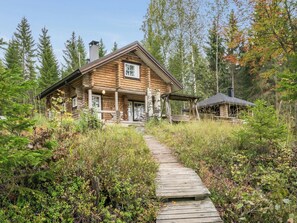 A log cabin with a wooden deck leading to the entrance, surrounded by dense forest.