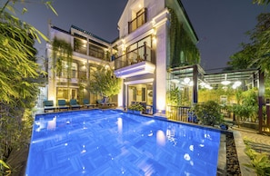 Un hotel de varios pisos con piscina, mobiliario exterior y abundante vegetación.