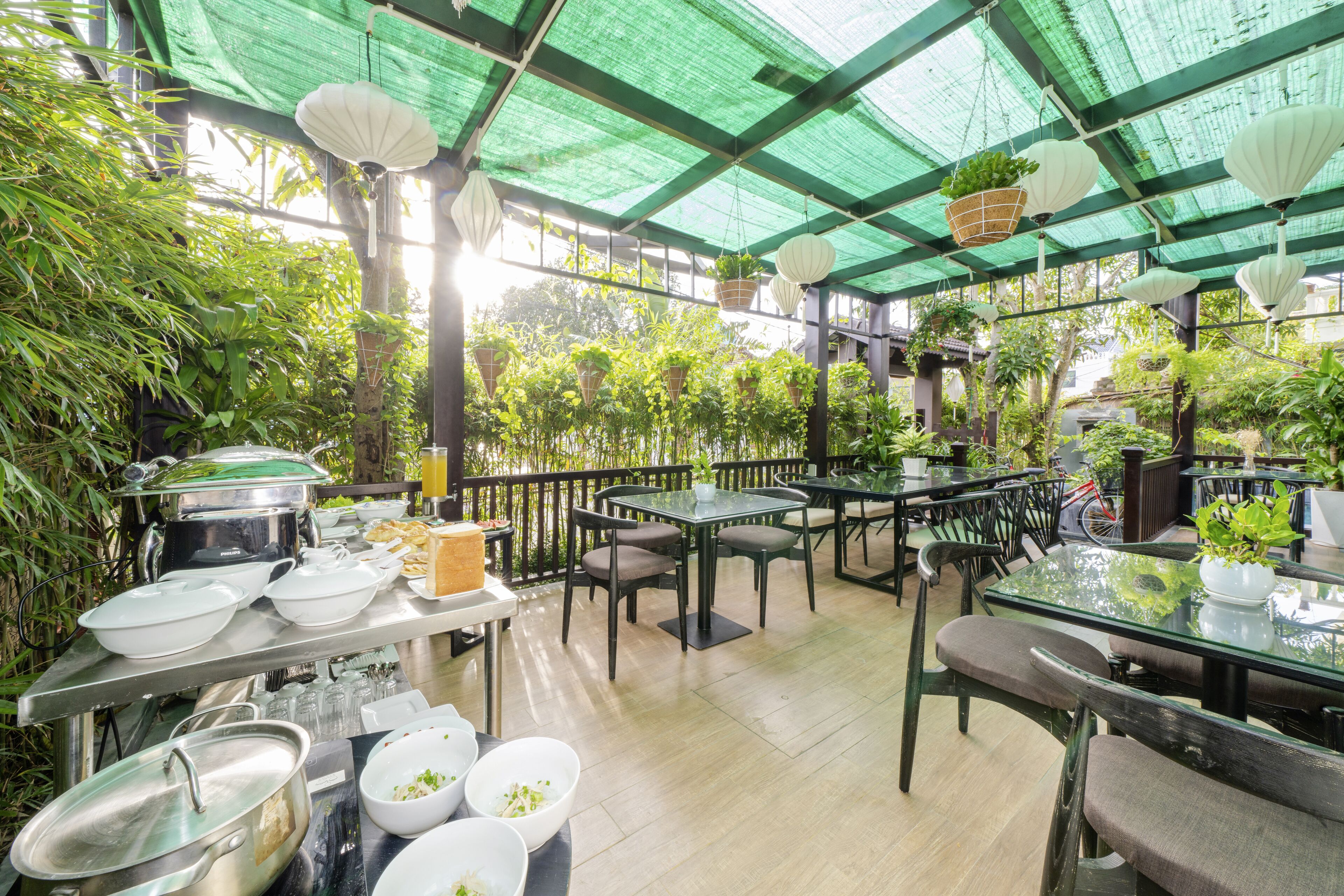 Un área de comedor al aire libre techada con mesas y sillas, rodeada de abundante vegetación y macetas colgantes.