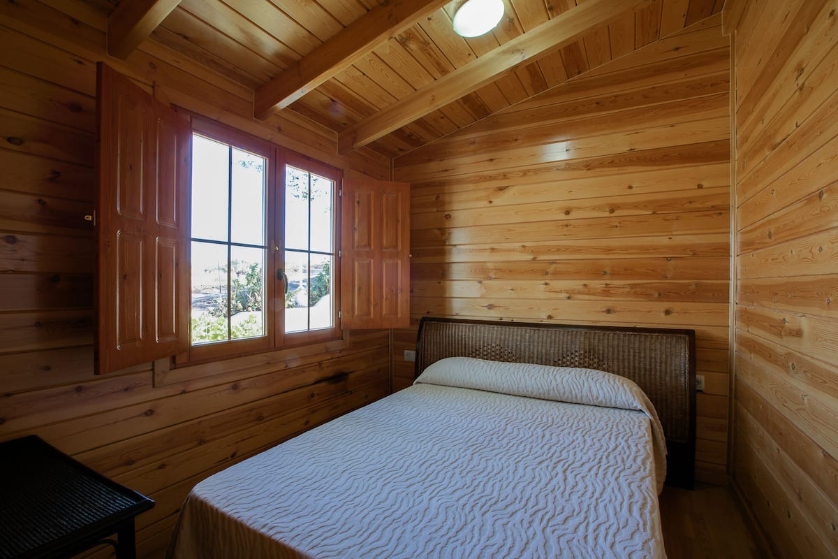 A wooden cabin bedroom with a bed, a window with shutters, and a ceiling light.