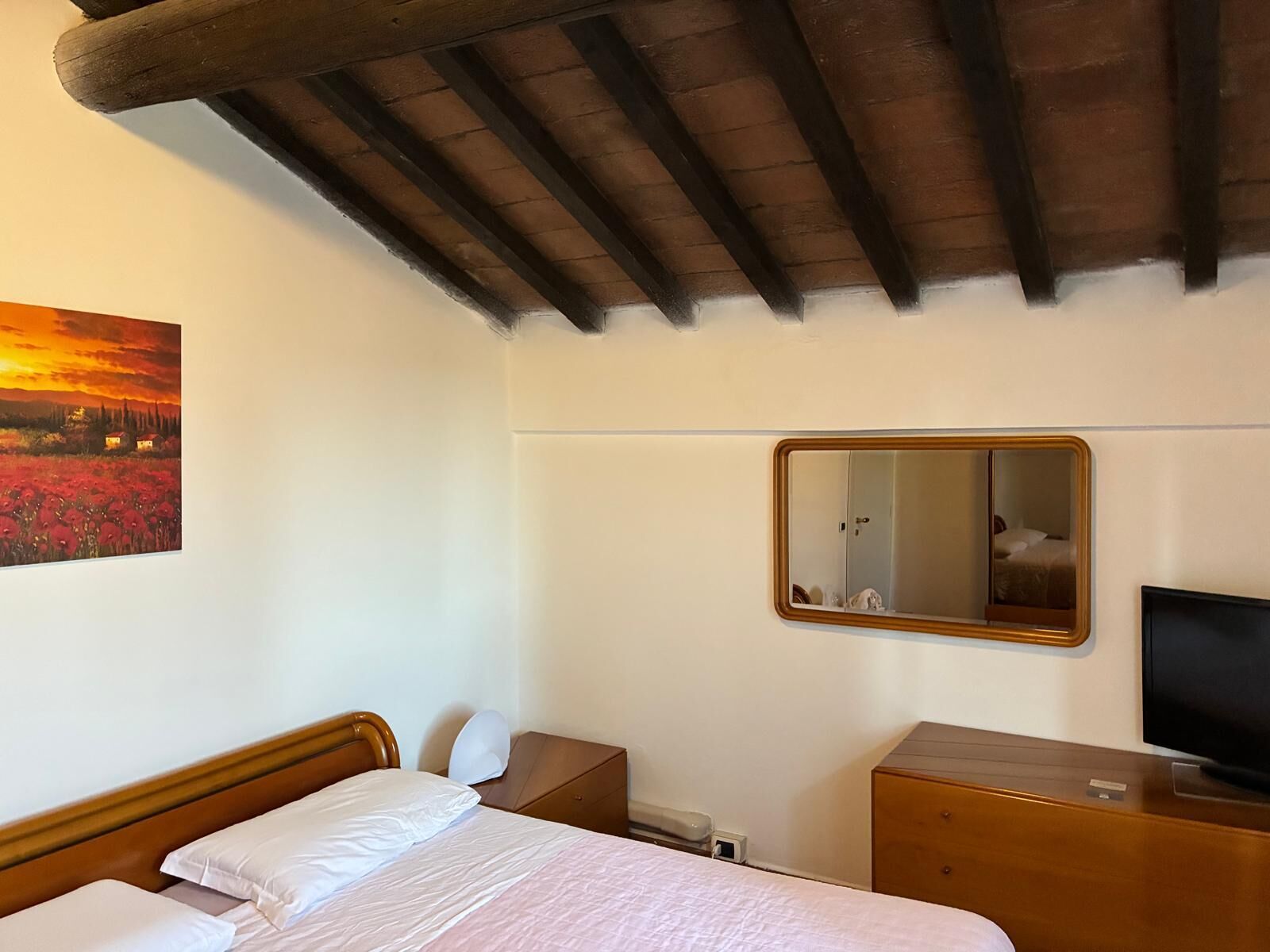 Une chambre d’hôtel avec un plafond en bois, un lit recouvert de draps blancs, une table de chevet avec une lampe, un téléviseur et un tableau représentant un champ de fleurs rouges.