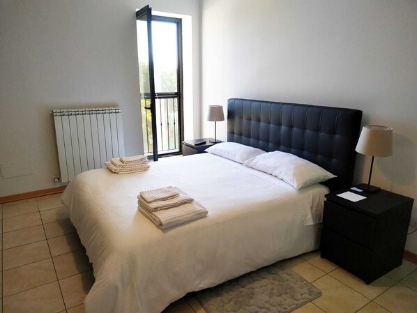 A neatly made bed with white linens and folded towels, a black headboard, a bedside table with a lamp, and a radiator on the wall.