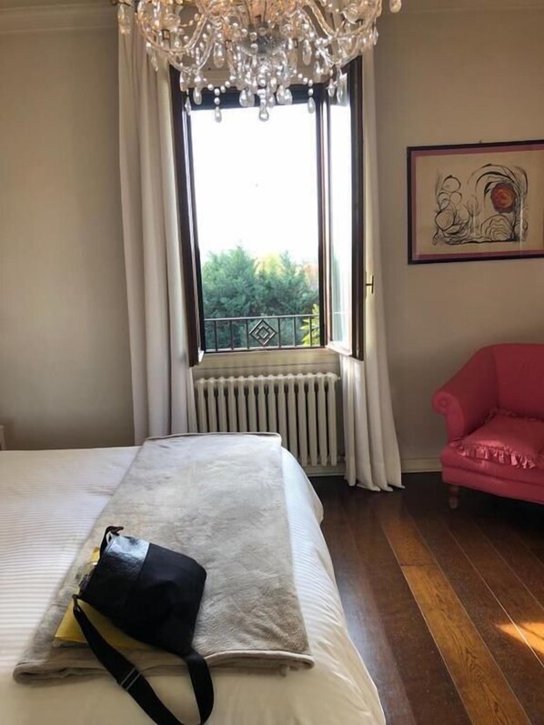 A hotel room with a bed, a pink armchair, a radiator, and a window with curtains. - B&B Villa delle Palme (Carpi)