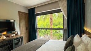 A hotel room with a bed, a TV, a desk, and a window with a view of greenery. - Falcon 1511 (Antalya)