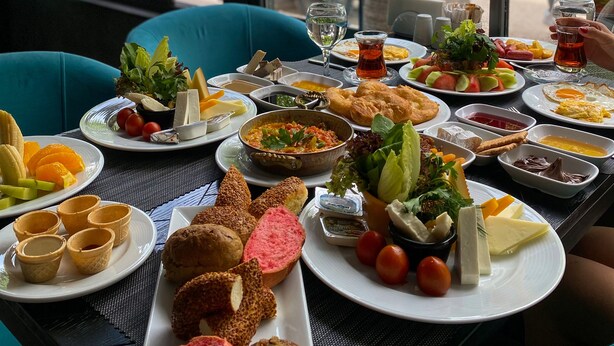 A table set with various dishes, including salads, bread, and desserts, in a room with large windows and teal chairs. - Falcon 1511 (Antalya)