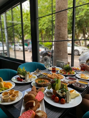 A table set with various dishes, including salads, bread, and desserts, in a room with large windows and teal chairs. - Falcon 1511 (Antalya)