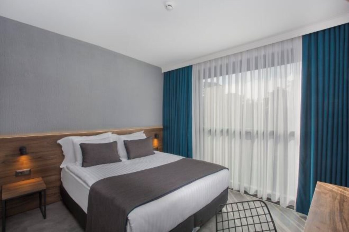 A hotel room with a bed, a nightstand, a window with curtains, and a view of the outdoors.