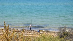 Une plage où deux personnes pêchent, avec une eau d’un bleu limpide et une végétation clairsemée.