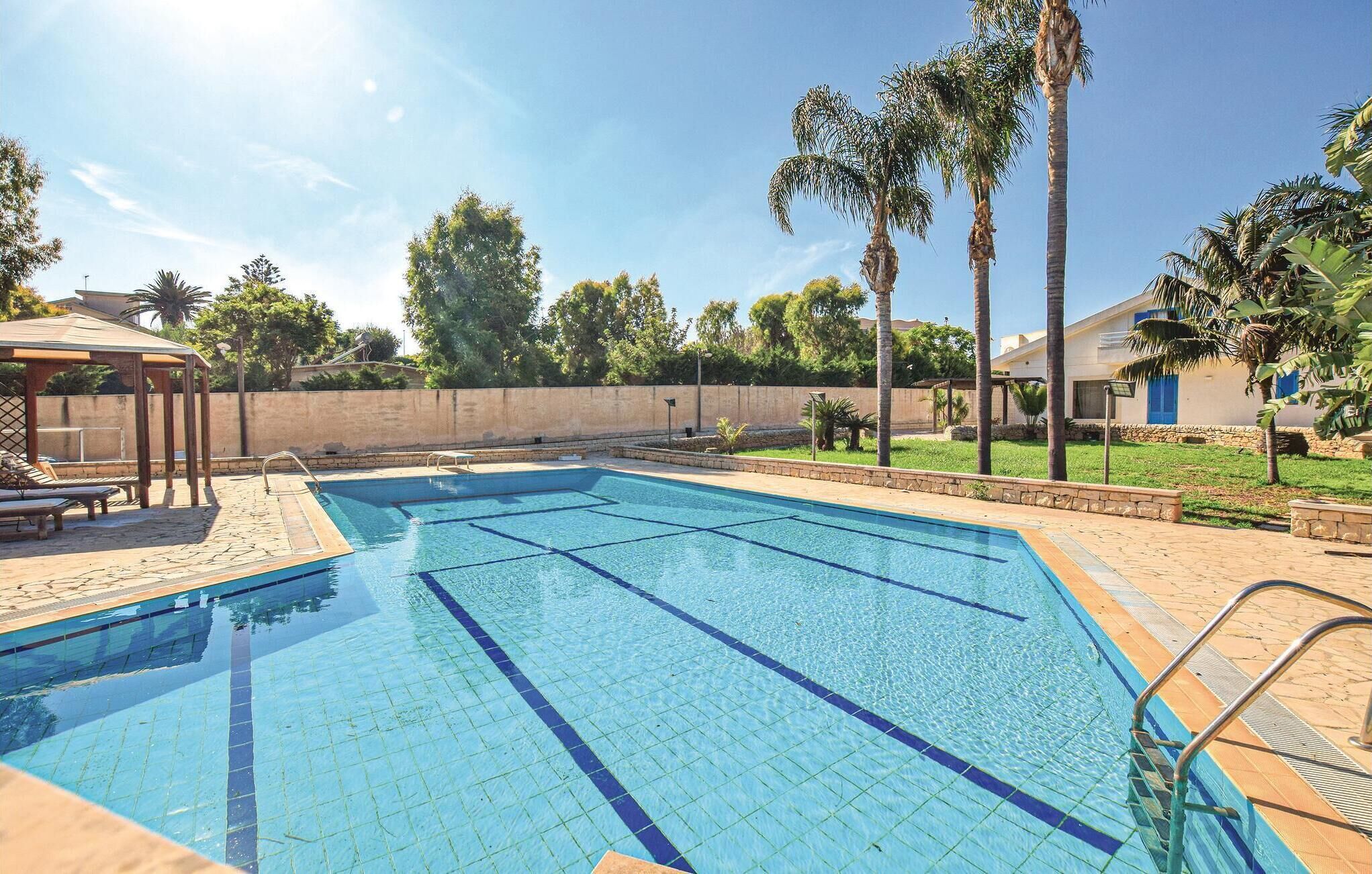 A swimming pool with clear blue water, surrounded by palm trees and a stone wall.