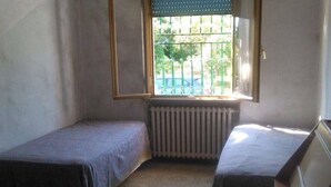 A small room with two single beds, a radiator, and a window with a view of a green fence and trees. - Holiday home in Val di Chiana (Castiglion Fiorentino)