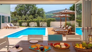 Un espace de restauration aménagé au bord de la piscine, avec une table dressée pour deux personnes, une piscine, des chaises longues et une vue sur le paysage environnant.