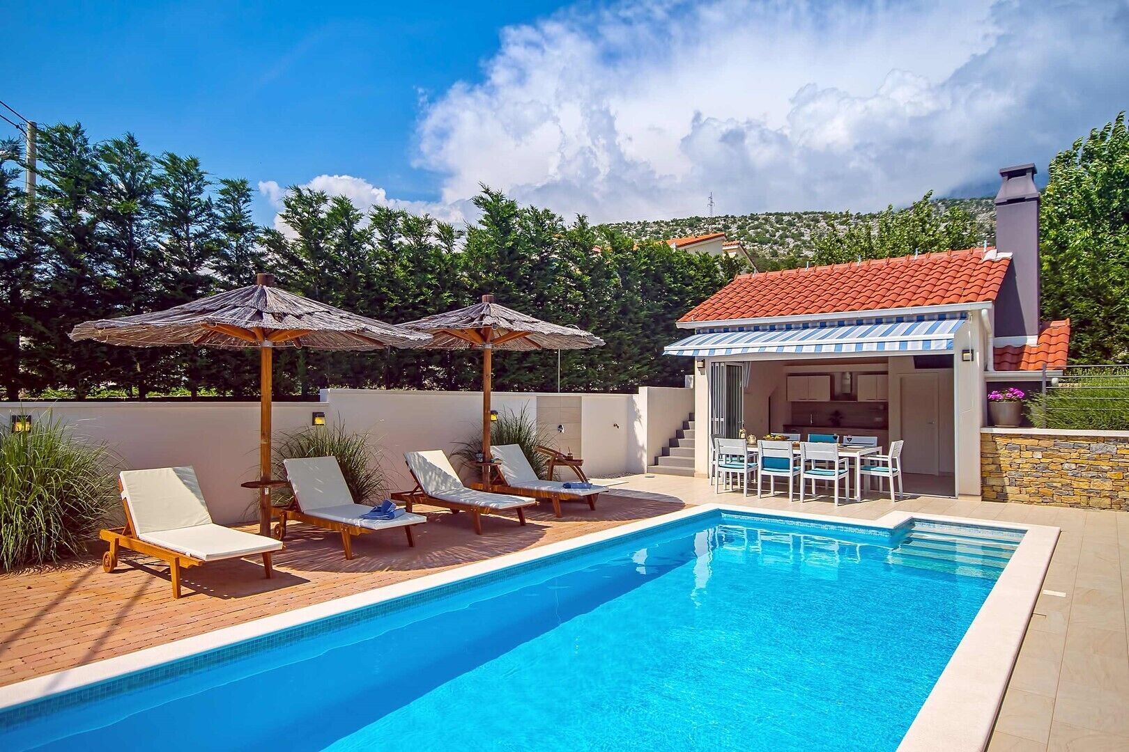 Une piscine avec des chaises longues et des parasols, un espace repas extérieur couvert et un mur en pierre.