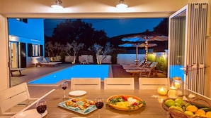 Un espace de restauration en plein air avec vue sur la piscine, doté de mobilier en bois et d’une table dressée avec des assiettes, des verres et un panier de fruits.