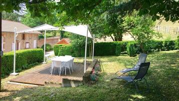 Une terrasse avec une table et des chaises, protégée par un auvent blanc, entourée d’un jardin bien entretenu et d’une haie.